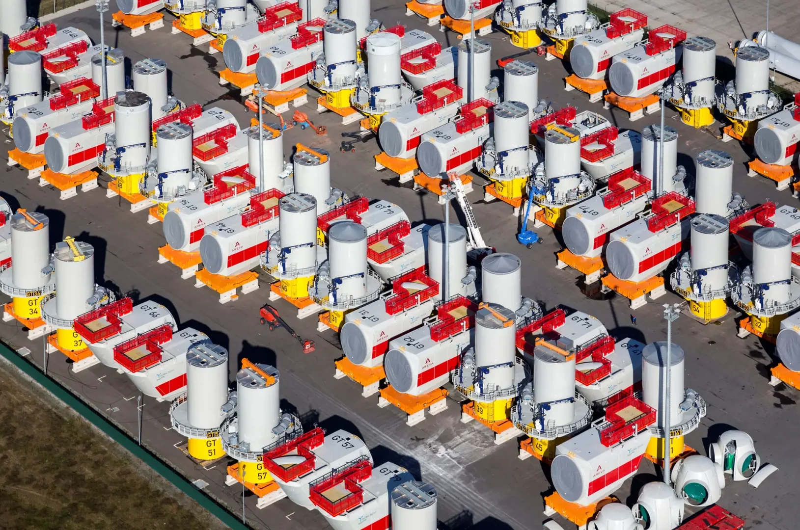Luftaufnahme eines Werftgeländes mit zahlreichen gelagerten Gondeln und Turmsegmenten für Offshore-Windkraftanlagen. Die Bauteile sind ordentlich aufgereiht und bereit für den Transport.
