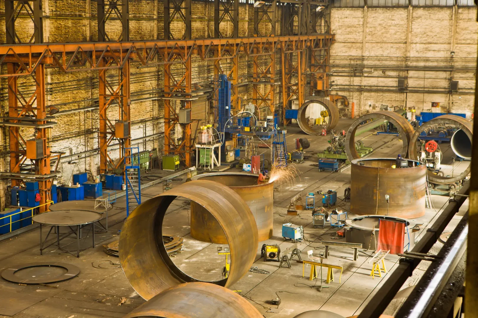 Innenaufnahme einer großen Industriehalle. Arbeiter schweißen an massive Stahlröhren für Offshore-Windkraftanlagen-Fundamente, Funkenflug ist sichtbar.