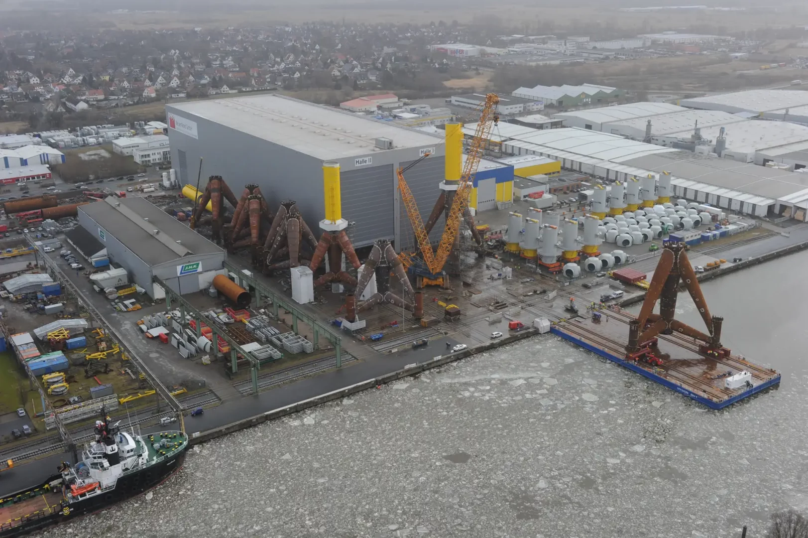 Luftaufnahme einer Offshore-Werft mit Produktionshallen, Kränen und Fundamentstrukturen. Am Kai liegen große Bauteile und ein Arbeitsschiff im Eiswasser.