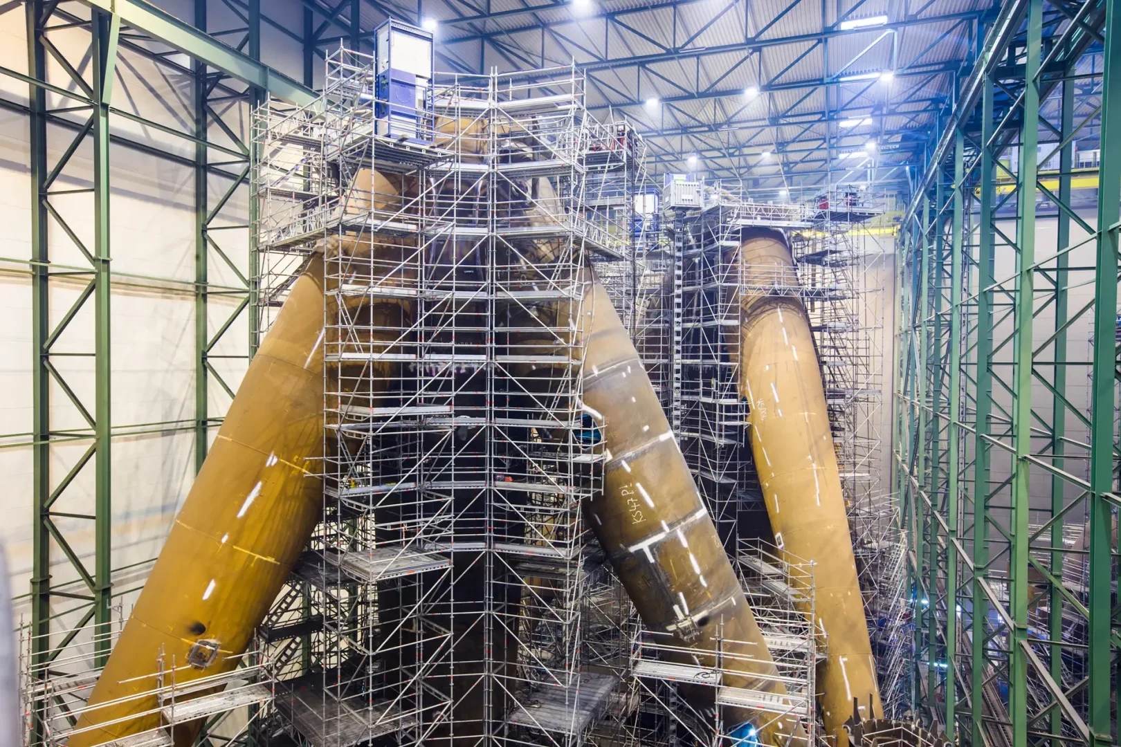 Innenaufnahme einer Werfthalle mit riesigen Stahlstrukturen für Offshore-Fundamente. Ein Gerüst umschließt die Bauteile für die Fertigung.