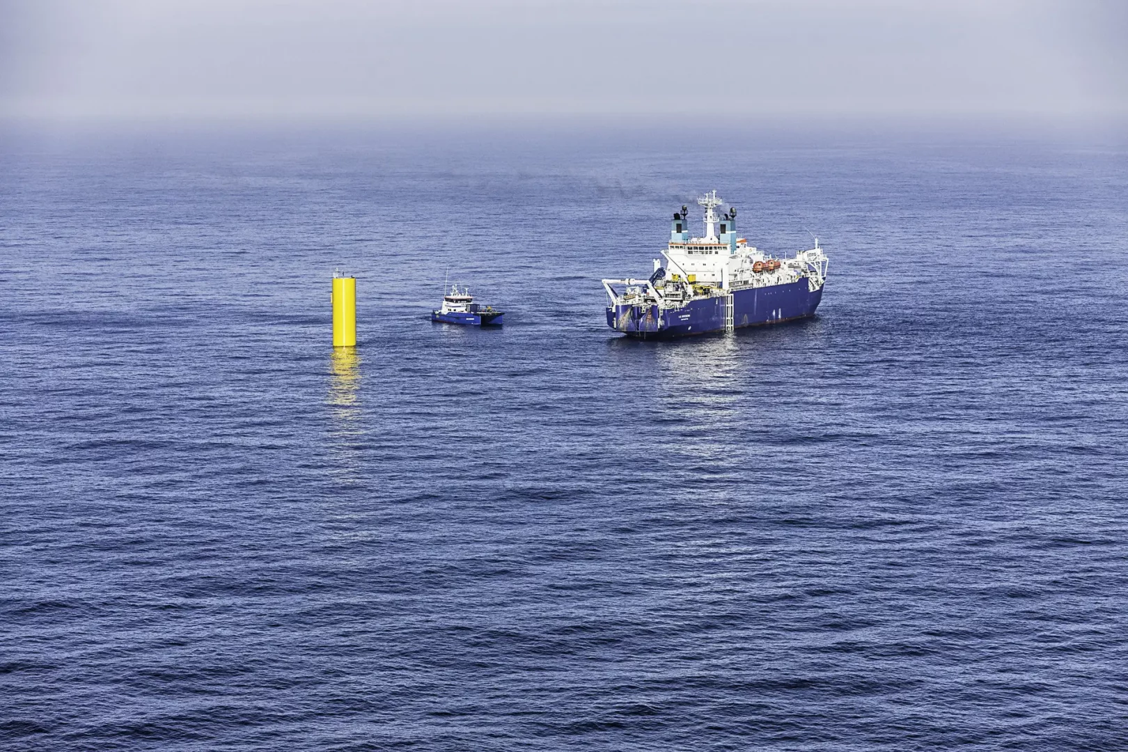 Ein Offshore-Spezialschiff liegt auf der Nordsee neben dem gelben Zentralrohr eines Tripod- Fundaments. Daneben ist ein kleineres Arbeitsschiff im Einsatz.