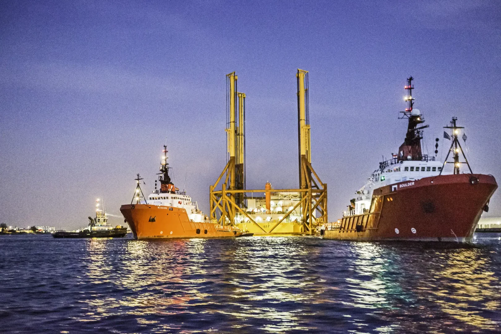 Nachtaufnahme von zwei roten Schleppschiffen beim Transport der großen Offshore-Umspannstation. Die beleuchteten Strukturen spiegeln sich auf der Wasseroberfläche.