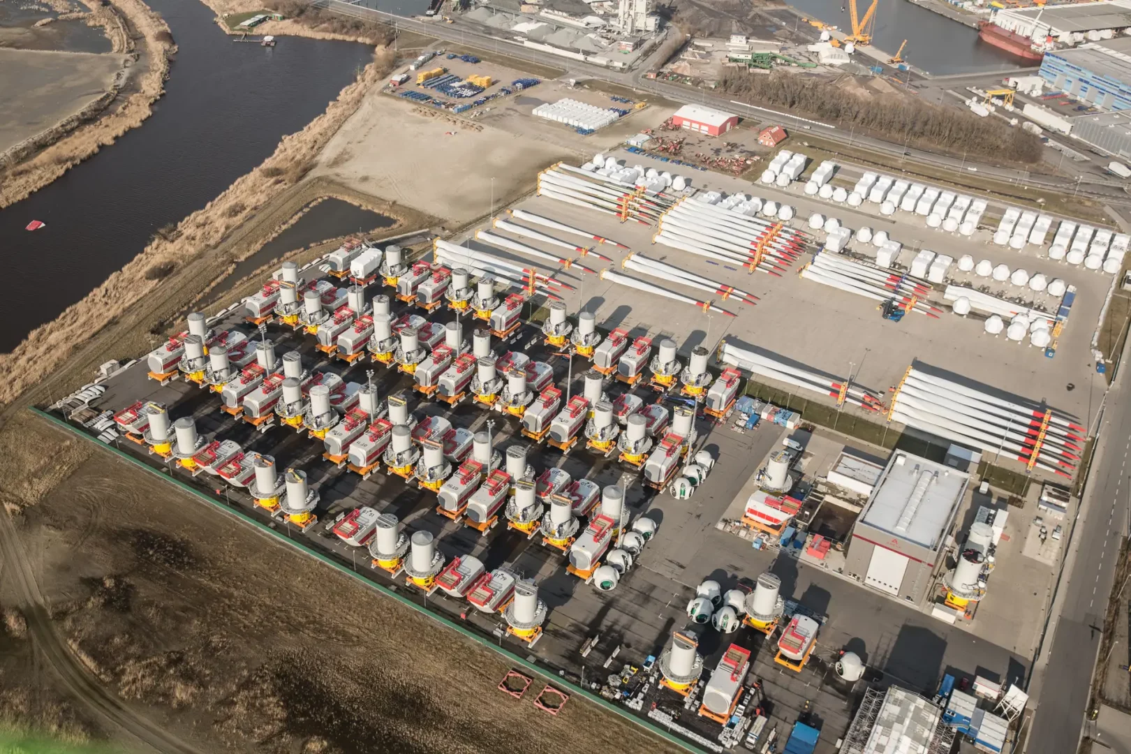 Luftaufnahme eines Werftgeländes mit zahlreichen gelagerten Gondeln, Rotorblättern und Turmsegmenten für Offshore-Windkraftanlagen. Die Komponenten sind für den Export und die Montage vorbereitet.
