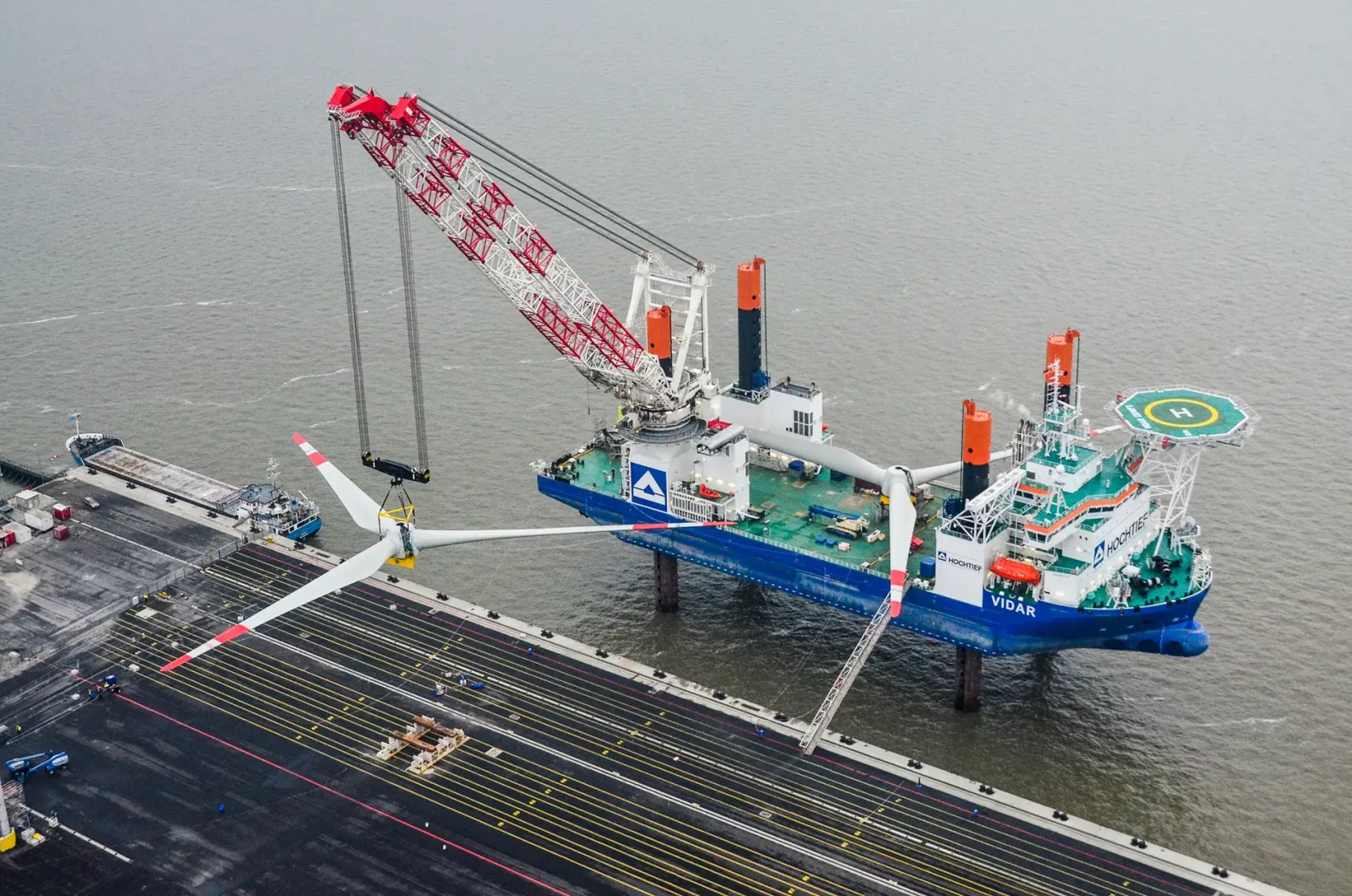 Ein Offshore-Installationsschiff mit großem Kran (HOCHTIEF Vidar) hebt einen Rotorstern vom Hafen auf das Schiff. Im Vordergrund die Kaianlage mit markierten Stellflächen.
