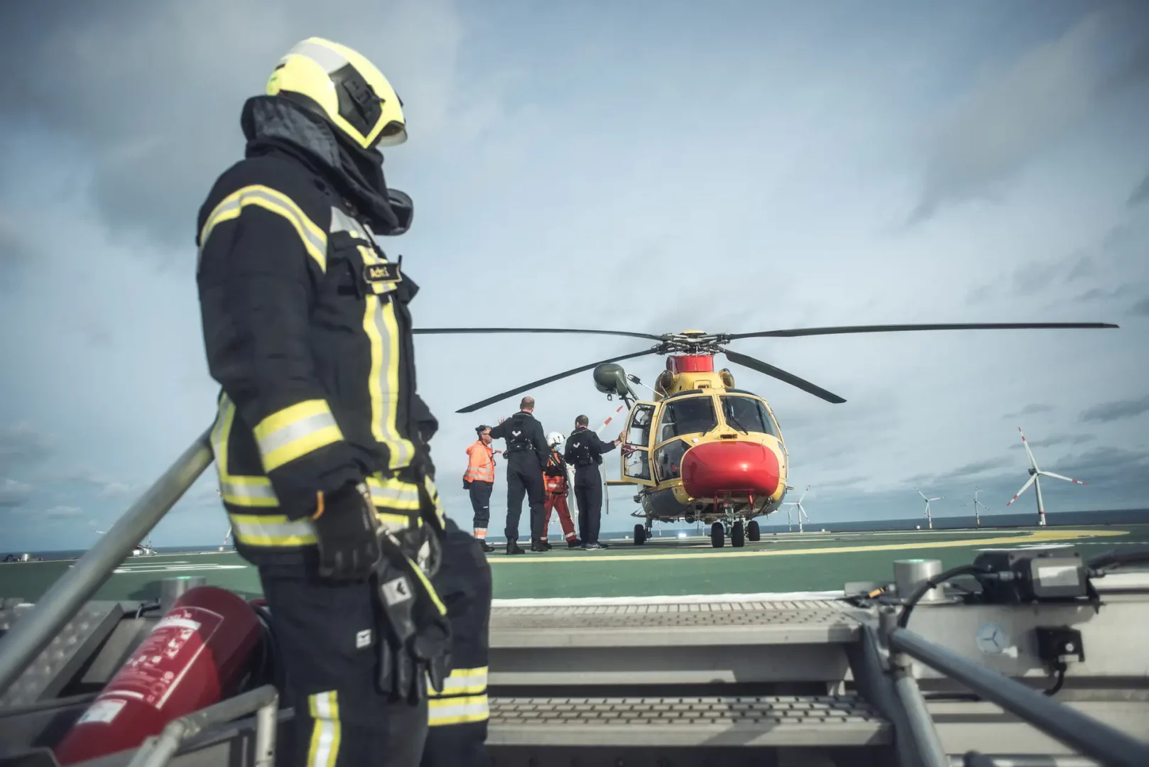 Übungseinsatz auf einer Offshore-Plattform: Feuerwehrkräfte und Sicherheitspersonal stehen vor einem gelben Rettungshubschrauber auf dem Helideck, im Hintergrund sind Windkraftanlagen zu sehen.