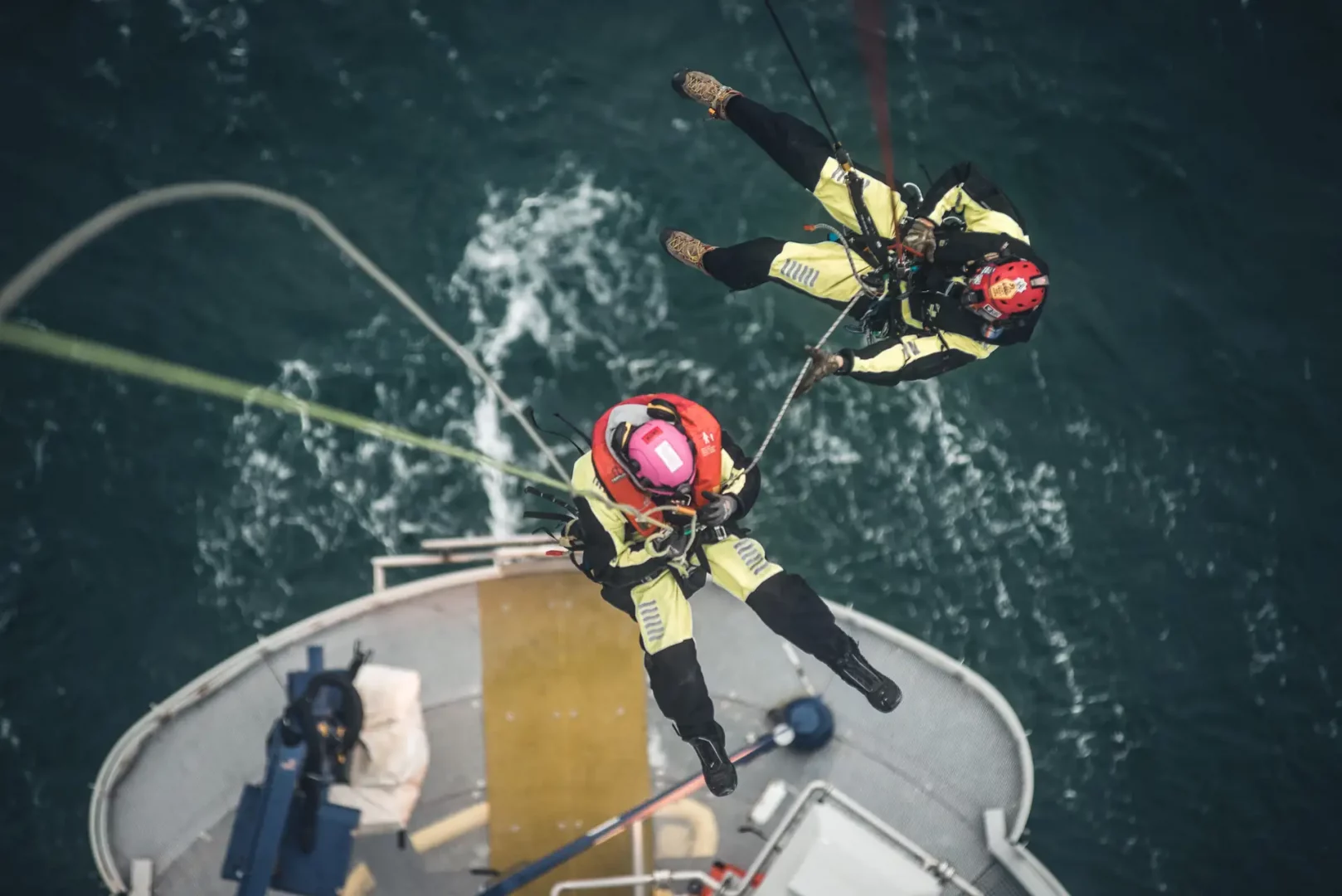 Rettungsübung über der Nordsee: Zwei Mitarbeitende in Schutzanzügen trainieren sich außen an der Windkraftanlage auf die untere Plattform abzuseilen.