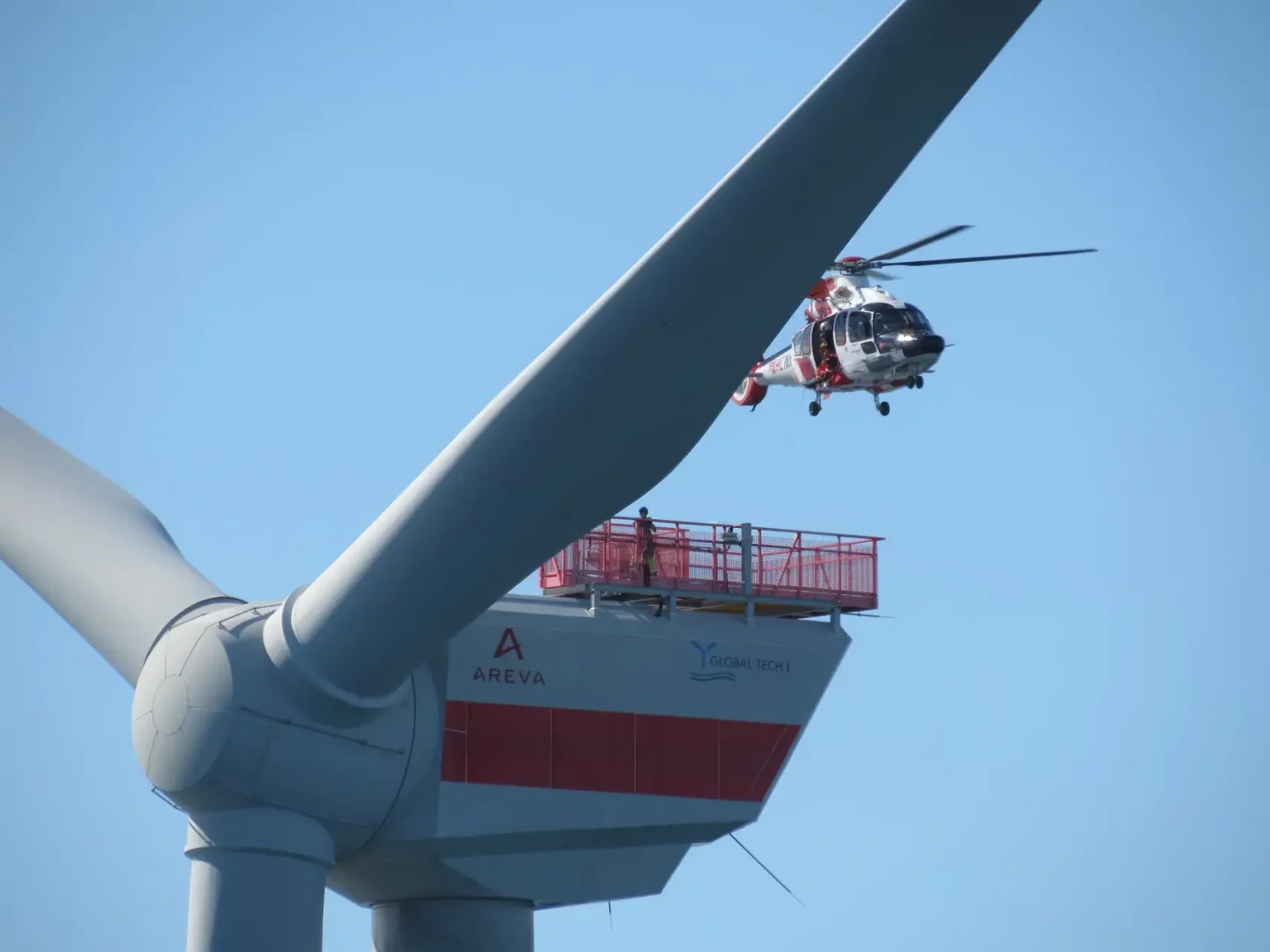 Ein Hubschrauber mit geöffneter Tür verweilt dicht über einer Windkraftanlage des Projekts Global Tech I. Deutlich sichtbar sind Gondel, Rotorblätter und Helihoist-Plattform.