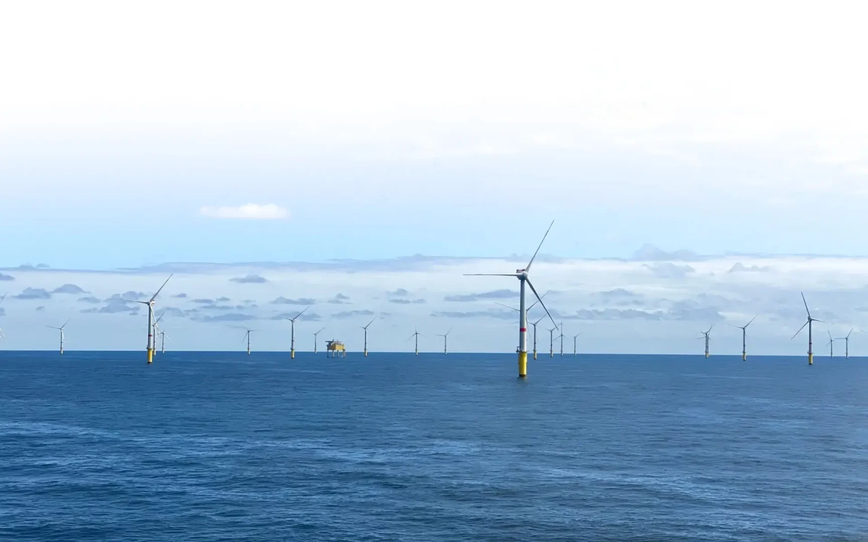 Panoramaaufnahme des Offshore-Windparks Global Tech I in der Nordsee. Zahlreiche Windkraftanlagen auf gelben Fundamenten sind über das Meer verteilt, im Hintergrund eine Offshore-Plattform.