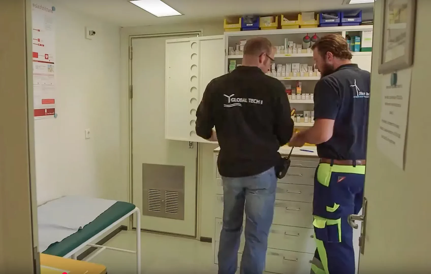 Medizinischer Raum auf einer Offshore-Plattform. Ein Mitarbeiter von Global Tech I und ein Medic besprechen sich vor einem Medikamentenschrank, links eine Untersuchungsliege.